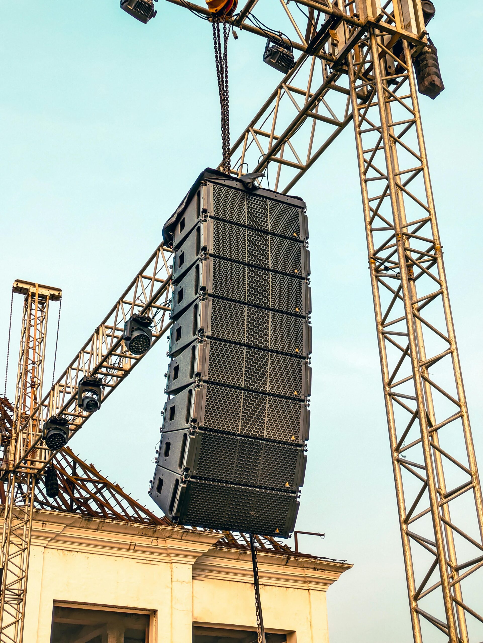 Massive speaker array suspended from crane system at urban construction site.